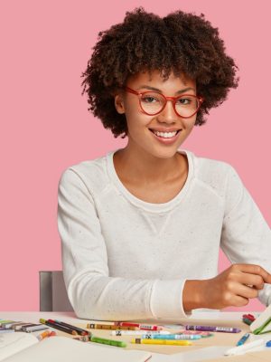 Horizontal shot of cheerful Afro African painter wears red rim spectacles, makes sketches in spiral notebook with pencil, enjoys aromatic beverage, surrounded with stationary, smiles positively reading and writing competitions for schools